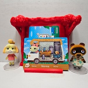 May include: A red plastic display stand holding a rectangular Animal Crossing Norma character card featuring a cow inside a camper van. The card is flanked by two small figurines: a yellow dog character on the left and a brown raccoon on the right.