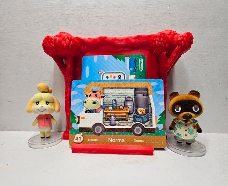 May include: A red plastic display stand holding a rectangular Animal Crossing Norma character card featuring a cow inside a camper van. The card is flanked by two small figurines: a yellow dog character on the left and a brown raccoon on the right.