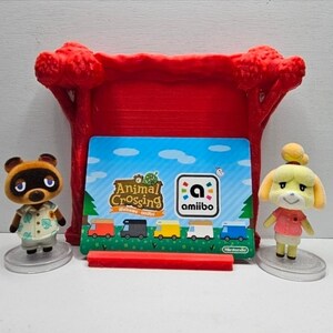May include: Two small Animal Crossing character figurines, one brown raccoon and one yellow dog, stand on clear circular bases flanking a rectangular Animal Crossing Welcome amiibo card displayed in a red plastic stand with tree-shaped sides.