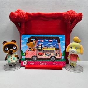 May include: Two small animal figurines standing on circular bases flanking a rectangular trading card. The card features a pink camper van illustration with the text 13, Kanga, Carrie, and Lola. A red plastic display stand sits behind the card.