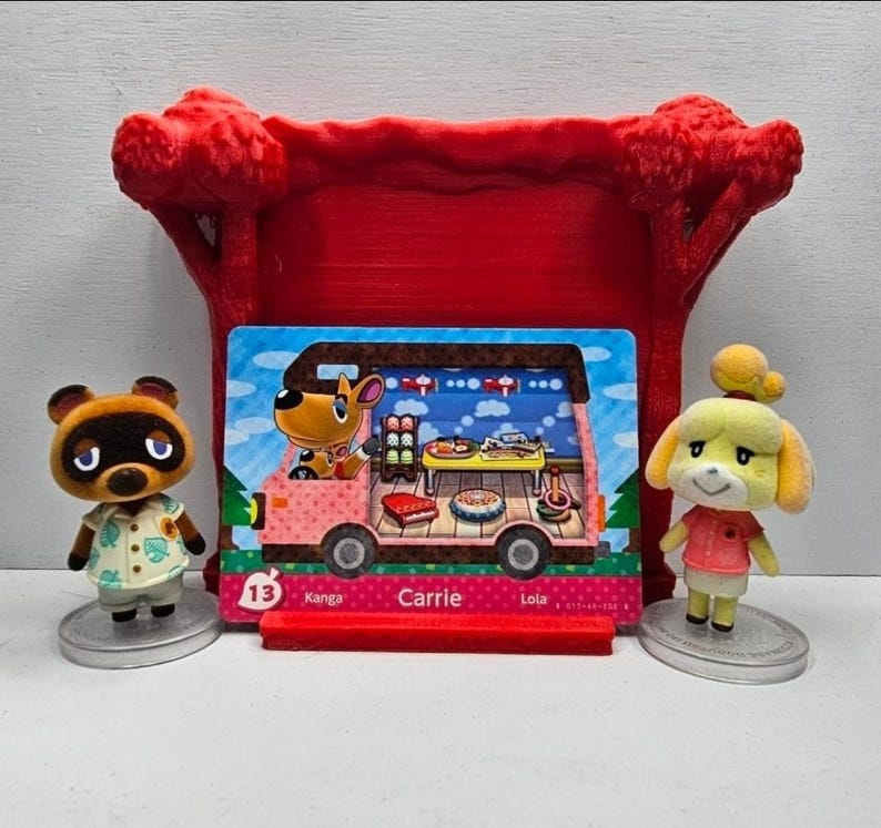 May include: Two small animal figurines standing on circular bases flanking a rectangular trading card. The card features a pink camper van illustration with the text 13, Kanga, Carrie, and Lola. A red plastic display stand sits behind the card.