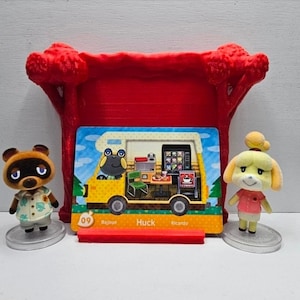 May include: Two Animal Crossing figurines, Tom Nook and Isabelle, stand on clear circular bases flanking a rectangular trading card. The card features a yellow camper van interior with the name Huck, number 09, and the text Bajoue and Ricardo.