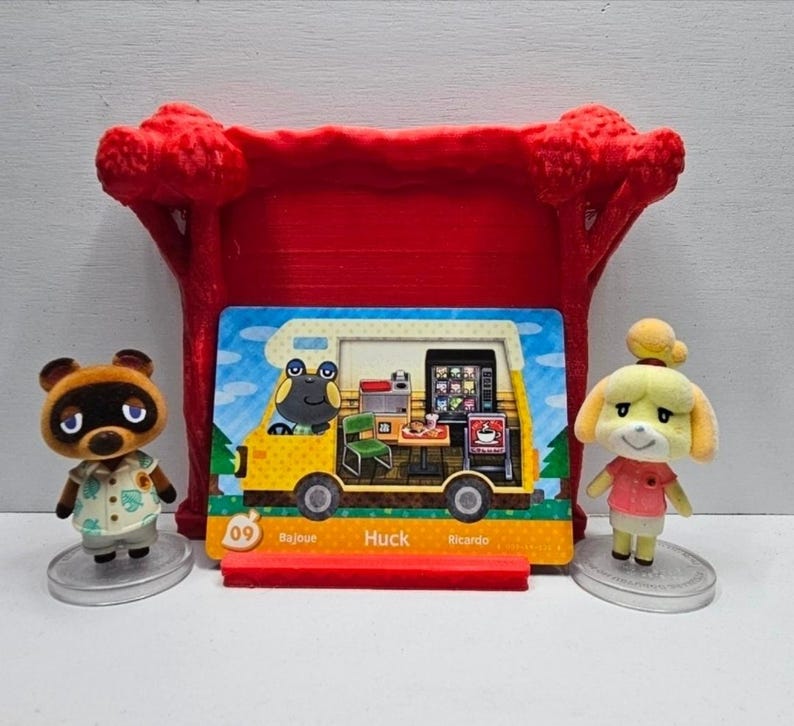 May include: Two Animal Crossing figurines, Tom Nook and Isabelle, stand on clear circular bases flanking a rectangular trading card. The card features a yellow camper van interior with the name Huck, number 09, and the text Bajoue and Ricardo.