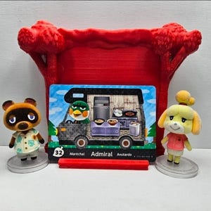 May include: Two small Animal Crossing character figurines, one brown raccoon and one yellow dog, stand beside a rectangular trading card featuring a green bird character inside a camper van. The card is displayed in a red 3D-printed stand.