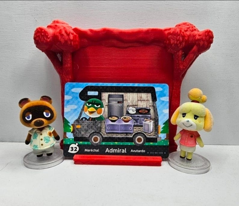 May include: Two small Animal Crossing character figurines, one brown raccoon and one yellow dog, stand beside a rectangular trading card featuring a green bird character inside a camper van. The card is displayed in a red 3D-printed stand.