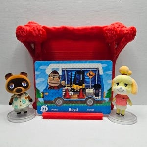 May include: Two small animal figurines, one brown raccoon and one yellow dog, stand on circular bases flanking a rectangular trading card. The card features a blue camper van with a gorilla character, a forklift, and the text 21 Primo Boyd Bunga.
