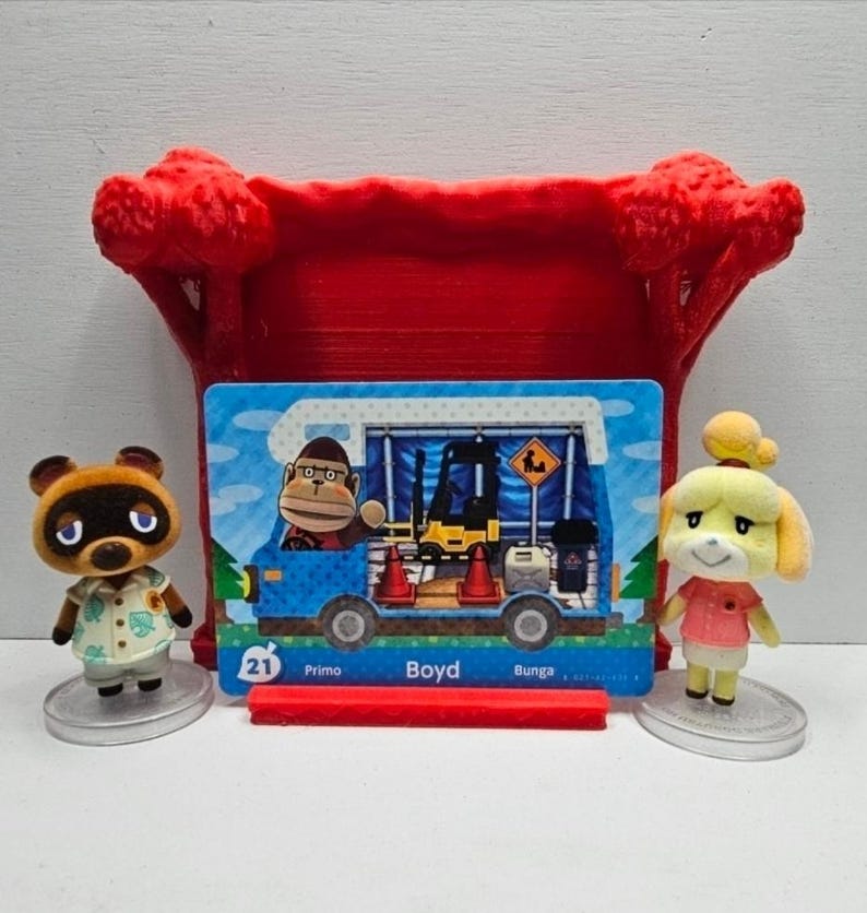 May include: Two small animal figurines, one brown raccoon and one yellow dog, stand on circular bases flanking a rectangular trading card. The card features a blue camper van with a gorilla character, a forklift, and the text 21 Primo Boyd Bunga.