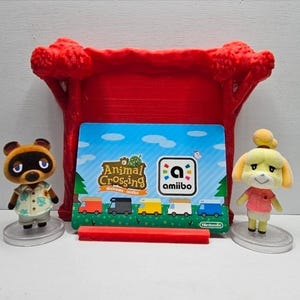 K&ouml;nnte beinhalten: Zwei kleine Animal Crossing-Figuren, ein brauner Waschb&auml;r und ein gelber Hund, stehen auf runden, transparenten Sockeln neben einer Animal Crossing Welcome amiibo-Karte, die in einem roten Kunststoffst&auml;nder mit baumf&ouml;rmigen Seiten steckt.