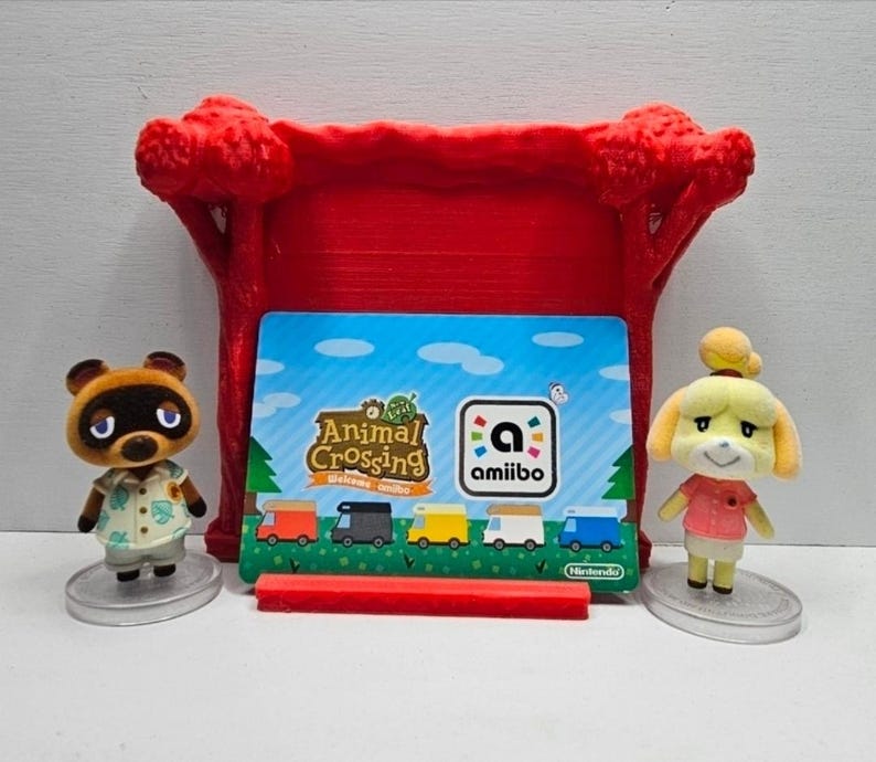 K&ouml;nnte beinhalten: Zwei kleine Animal Crossing-Figuren, ein brauner Waschb&auml;r und ein gelber Hund, stehen auf runden, transparenten Sockeln neben einer Animal Crossing Welcome amiibo-Karte, die in einem roten Kunststoffst&auml;nder mit baumf&ouml;rmigen Seiten steckt.