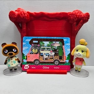 K&ouml;nnte beinhalten: Ein roter 3D-gedruckter Pr&auml;sentationsst&auml;nder mit einer Animal Crossing Amiibo-Karte, die Olive in einem rosa Wohnmobil zeigt. Die Karte tr&auml;gt die Nummer 45 und den Namen Olive. Zwei kleine Animal Crossing-Figuren stehen auf runden Sockeln.