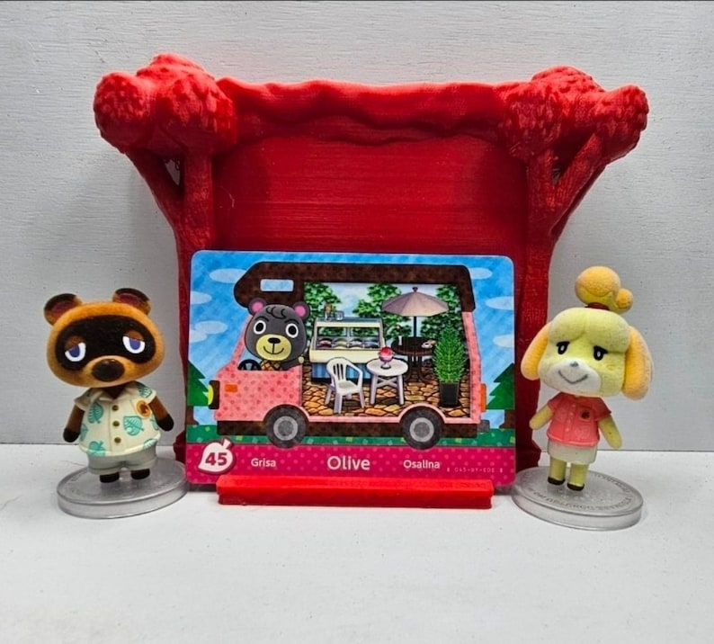 K&ouml;nnte beinhalten: Ein roter 3D-gedruckter Pr&auml;sentationsst&auml;nder mit einer Animal Crossing Amiibo-Karte, die Olive in einem rosa Wohnmobil zeigt. Die Karte tr&auml;gt die Nummer 45 und den Namen Olive. Zwei kleine Animal Crossing-Figuren stehen auf runden Sockeln.