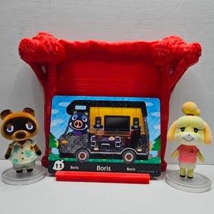 May include: Two small animal figurines, one brown raccoon and one yellow dog, stand beside a rectangular trading card featuring a black camper van with the name Boris printed on it. A red plastic tree-shaped display stand sits behind the card.