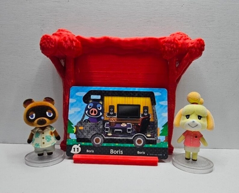 May include: Two small animal figurines, one brown raccoon and one yellow dog, stand beside a rectangular trading card featuring a black camper van with the name Boris printed on it. A red plastic tree-shaped display stand sits behind the card.