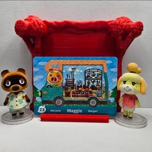 May include: A rectangular Animal Crossing trading card featuring Maggie the pig inside a teal camper van, displayed in a red plastic stand between two small animal figurines, one a brown raccoon and the other a yellow dog, both on clear bases.