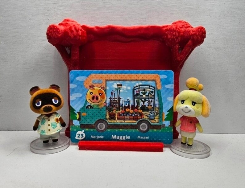 May include: A rectangular Animal Crossing trading card featuring Maggie the pig inside a teal camper van, displayed in a red plastic stand between two small animal figurines, one a brown raccoon and the other a yellow dog, both on clear bases.