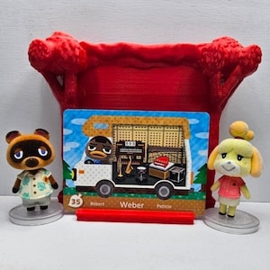 May include: Two small Animal Crossing figurines, Tom Nook and Isabelle, stand on circular bases flanking a rectangular trading card featuring a camper van with the character Weber, labeled with the number 35, all set before a red plastic tree frame.