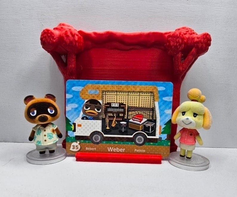May include: Two small Animal Crossing figurines, Tom Nook and Isabelle, stand on circular bases flanking a rectangular trading card featuring a camper van with the character Weber, labeled with the number 35, all set before a red plastic tree frame.