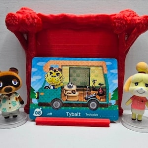 May include: Two small flocked animal figurines stand beside a rectangular trading card featuring a teal camper van with a tiger character inside. The card displays the text 08 Jeff Tybalt Teobaldo. A red plastic stand supports the card.