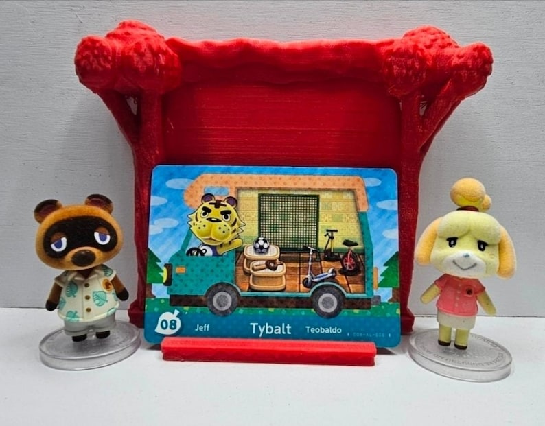 May include: Two small flocked animal figurines stand beside a rectangular trading card featuring a teal camper van with a tiger character inside. The card displays the text 08 Jeff Tybalt Teobaldo. A red plastic stand supports the card.