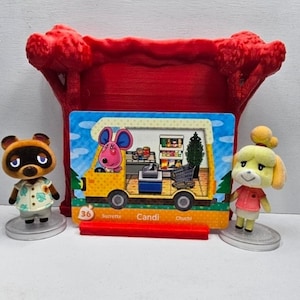 May include: A red 3D-printed display stand holding a rectangular Animal Crossing trading card featuring a yellow camper van with a pink mouse character inside, flanked by two small animal figurines on circular bases, Tom Nook and Isabelle.