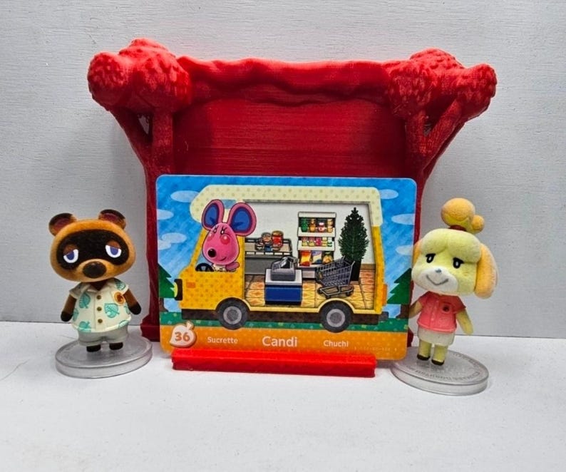 May include: A red 3D-printed display stand holding a rectangular Animal Crossing trading card featuring a yellow camper van with a pink mouse character inside, flanked by two small animal figurines on circular bases, Tom Nook and Isabelle.