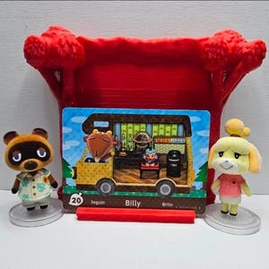 May include: A red plastic display stand holding an Animal Crossing Billy character card numbered 20, flanked by two small figurines of Tom Nook and Isabelle on clear circular bases. The card depicts a yellow camper van interior with a goat character.