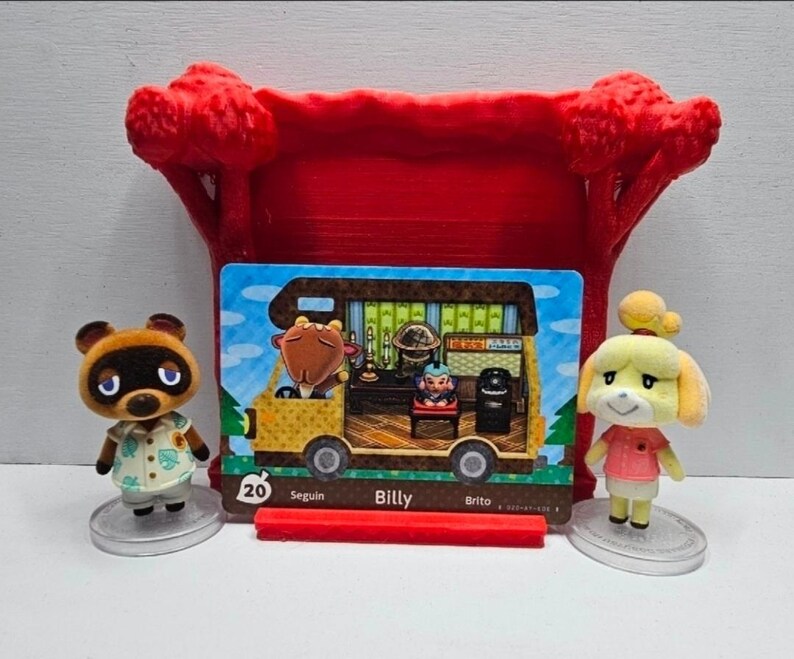 May include: A red plastic display stand holding an Animal Crossing Billy character card numbered 20, flanked by two small figurines of Tom Nook and Isabelle on clear circular bases. The card depicts a yellow camper van interior with a goat character.