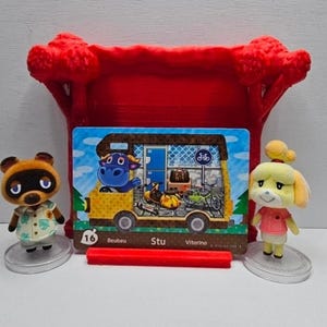 May include: A rectangular Animal Crossing trading card featuring a yellow camper van with a blue bull character, parked in front of a red 3D-printed tree stand. Two small figurines, a brown raccoon and a yellow dog, stand on clear circular bases.