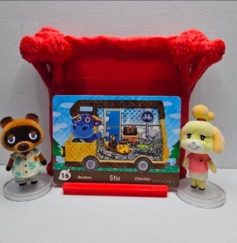 May include: A rectangular Animal Crossing trading card featuring a yellow camper van with a blue bull character, parked in front of a red 3D-printed tree stand. Two small figurines, a brown raccoon and a yellow dog, stand on clear circular bases.