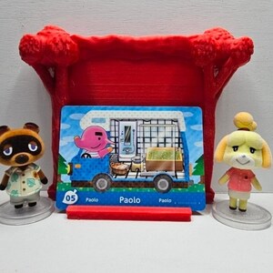 May include: Two small animal figurines, one brown raccoon and one yellow dog, stand on circular bases flanking a rectangular trading card. The card features a pink elephant in a blue camper van with the text 05 Paolo. A red plastic stand sits behind.