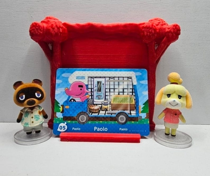 May include: Two small animal figurines, one brown raccoon and one yellow dog, stand on circular bases flanking a rectangular trading card. The card features a pink elephant in a blue camper van with the text 05 Paolo. A red plastic stand sits behind.