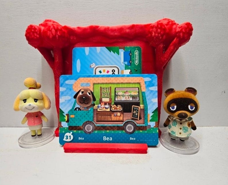May include: A red 3D-printed display stand holding an Animal Crossing Bea character card, featuring a brown dog inside a camper van. Two small flocked figurines of Isabelle and Tom Nook stand on clear circular bases on either side of the display.