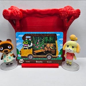 May include: A rectangular Animal Crossing trading card featuring a yellow camper van with a green alligator character named Boots, displayed in a red 3D-printed stand between two small figurines of Tom Nook and Isabelle on clear circular bases.