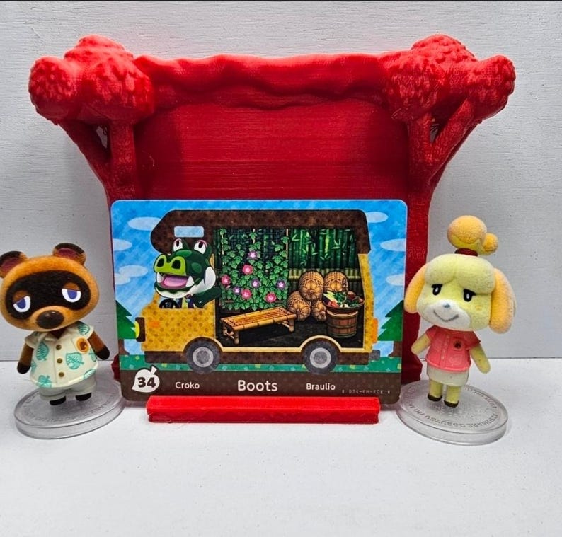 May include: A rectangular Animal Crossing trading card featuring a yellow camper van with a green alligator character named Boots, displayed in a red 3D-printed stand between two small figurines of Tom Nook and Isabelle on clear circular bases.