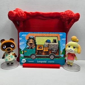 May include: A small brown raccoon figurine in a light green leaf-print shirt, a yellow dog figurine in a pink shirt, and a rectangular trading card featuring a green camper van with the name Leopold, all displayed before a red plastic tree stand.