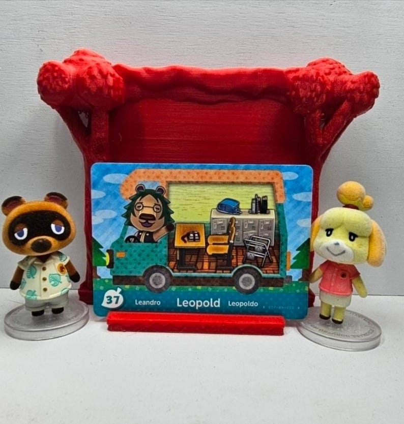 May include: A small brown raccoon figurine in a light green leaf-print shirt, a yellow dog figurine in a pink shirt, and a rectangular trading card featuring a green camper van with the name Leopold, all displayed before a red plastic tree stand.