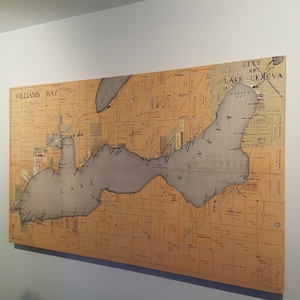 May include: A vintage map of Lake Geneva, Wisconsin, with the text "City of Lake Geneva" and "Williams Bay" printed on the map. The map is printed on a beige background with brown lines and text. The lake is shaded in gray.
