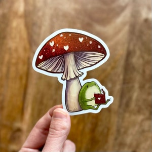 May include: A red and white mushroom with white hearts on the cap, a green frog sitting under the mushroom reading a red book with a white heart on the cover.