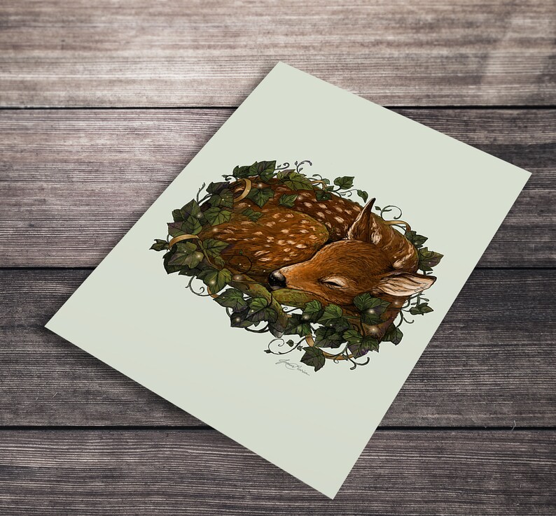 May include: A watercolor illustration of a sleeping fawn curled up in a wreath of green ivy with white flowers. The fawn has brown and white spots and closed eyes.