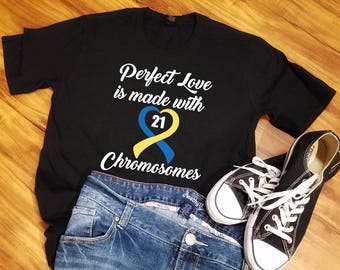 May include: Black t-shirt with white text that reads "Perfect Love is made with 21 Chromosomes". A yellow and blue ribbon with a heart and the number 21 is in the center of the text. The t-shirt is laying on a wooden surface with blue jeans and black sneakers.