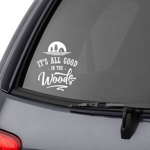 Könnte beinhalten: Ein grauer SUV mit einem weißen Vinyl-Aufkleber auf der Heckscheibe, auf dem "It's all good in the Woods" steht. Der Aufkleber zeigt eine Silhouette von Bäumen und einem Lagerfeuer.
