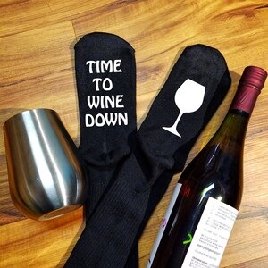 May include: A pair of black socks with white text that reads "Time to Wine Down" and a white wine glass graphic. A bottle of wine and a stainless steel wine glass are also included.