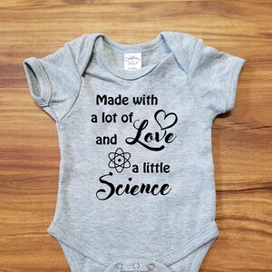 Könnte beinhalten: Grauer Baby-Strampler mit schwarzem Text, der "Made with a lot of Love and a little Science" lautet. Der Text enthält ein Herz und ein Atom-Symbol.