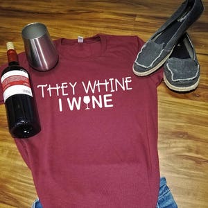 K&ouml;nnte beinhalten: Ein maroonfarbenes T-Shirt mit dem Text "They whine I wine" und einer Grafik eines Weinglases. Das Shirt liegt auf einer blauen Jeans und einer Holzoberfl&auml;che. Auf der Oberfl&auml;che befinden sich eine Weinflasche und ein silbernes Weinglas.
