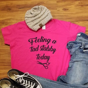 Op de afbeelding: Een casual outfit met een felroze t-shirt met de tekst "Feeling a tad stabby today" en een schaargrafiek. Inclusief een grijze gebreide beanie, een blauwe jeans en zwarte Converse sneakers met witte veters.