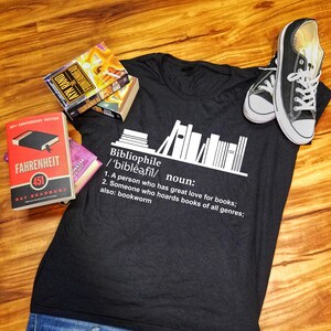 May include: A black t-shirt with a white graphic of books on a shelf and the definition of the word "bibliophile". The text reads: "Bibliophile /'biblēǝ,fil/ noun: 1. A person who has great love for books; 2. Someone who hoards books of all genres; also: bookworm"