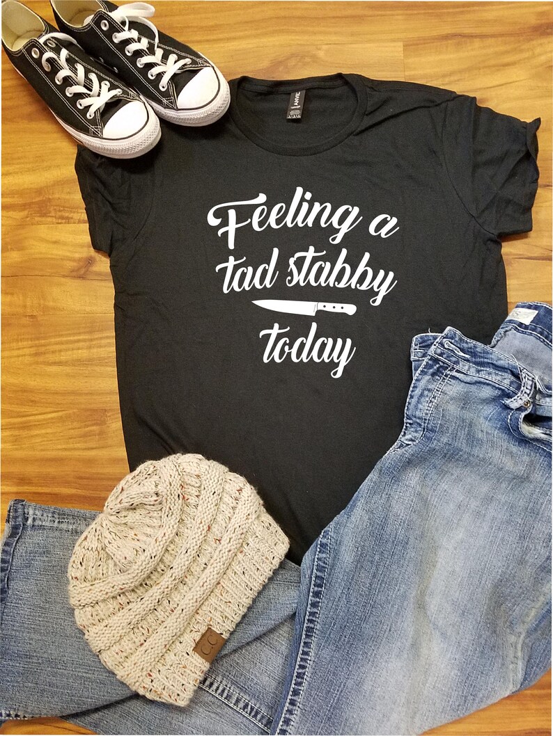Op de afbeelding: Een zwart t-shirt met de tekst "Feeling a tad stabby today" en een mesafbeelding. Het shirt wordt gecombineerd met een blauwe spijkerbroek, een beige gebreide muts en zwarte canvas sneakers.