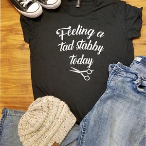 Op de afbeelding: Zwart t-shirt met de tekst "Feeling a tad stabby today" en een schaargrafiek. Inclusief zwarte sneakers, een lichtblauwe jeans en een beige gebreide muts.