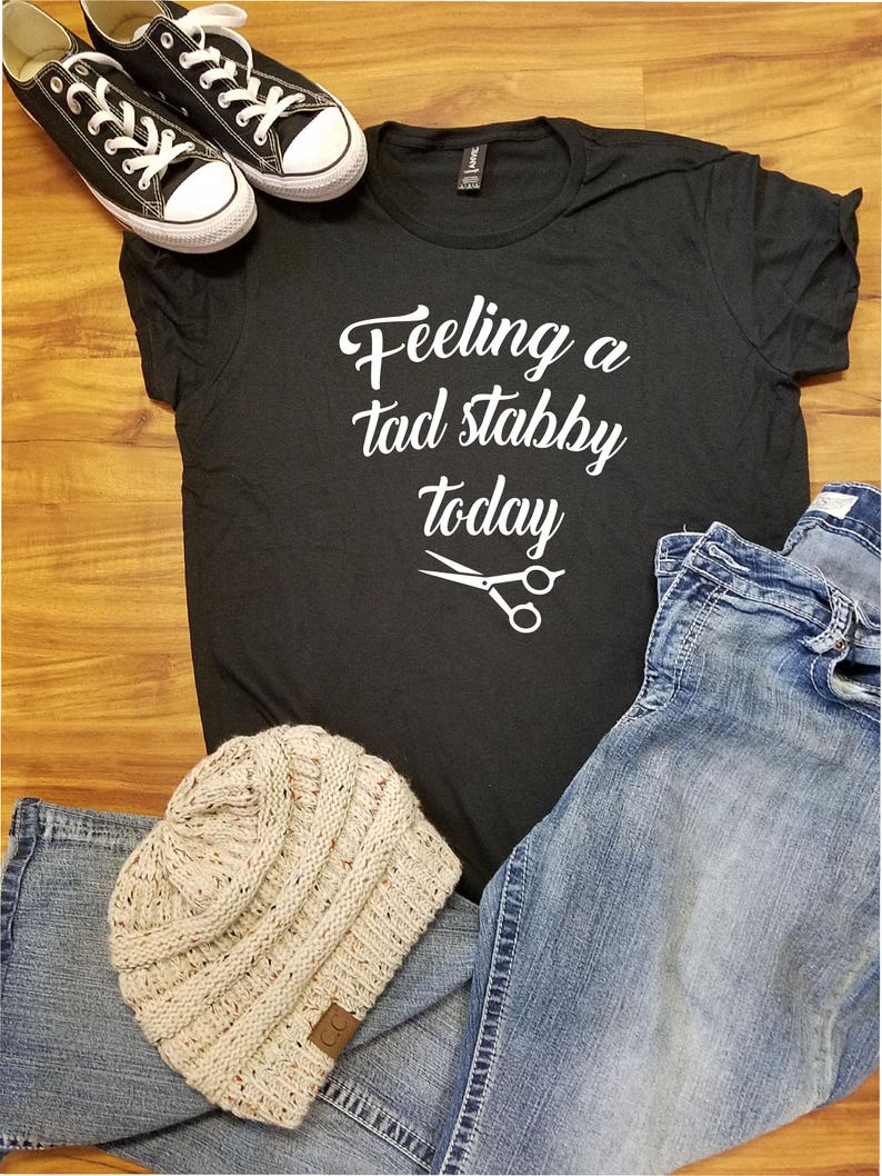Op de afbeelding: Zwart t-shirt met de tekst "Feeling a tad stabby today" en een schaargrafiek. Inclusief zwarte sneakers, een lichtblauwe jeans en een beige gebreide muts.