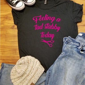 Op de afbeelding: Een casual ensemble met een zwart t-shirt met de roze tekst "Feeling a tad stabby today", gecombineerd met een blauwe jeans, een beige gebreide muts en zwarte canvas sneakers met witte veters.